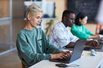 Side view of young classmates networking with focus on blond Asian guy typing on laptop keyboard while looking at screen and decoding data