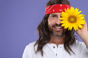 Stylish hippie man covering eye with sunflower on violet background, space for text