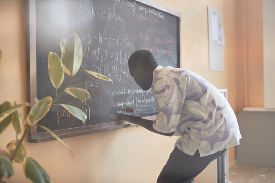 Young male student with tablet bending in front of blackboard while writing down formulas with chalk before explaining new subject