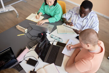 Above angle of intercultural students making notes and using mobile gadgets while sitting by large table in classroom and working at lesson