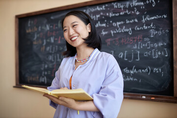 Happy young Asian female teacher with open book looking at students while standing by blackboard with formulas and explaining new subject