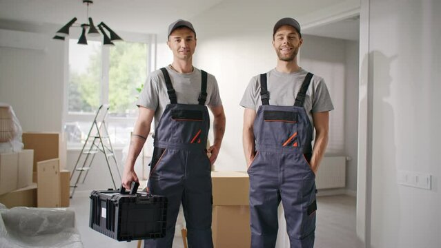 Repair masters finished apartment update. Male contractors in overalls satisfied with final result. Two repairmen smiling at camera. Craftsman posing with hands in pockets. Man holding tools box.
