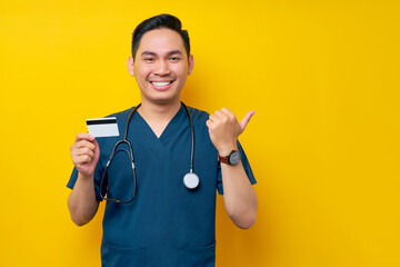 Smiling cheerful young Asian male doctor or nurse wearing blue uniform holding credit bank card and pointing finger aside workspace isolated on yellow background. Healthcare medicine concept - obrazy, fototapety, plakaty