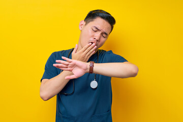 Bored young Asian male doctor or nurse wearing blue uniform looking at his watch to check time and yawning tired covering his mouth with a hand isolated on yellow background
