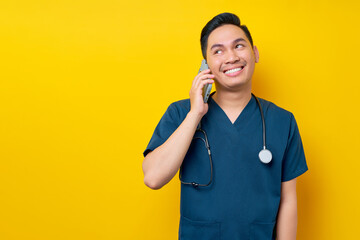 Smiling satisfied young Asian male doctor or nurse wearing blue uniform talking on mobile phone...