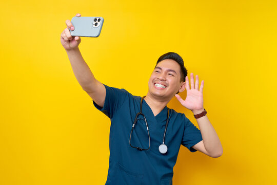 Smiling happy young Asian male doctor or nurse wearing blue uniform with a smartphone making video call or taking selfie isolated on yellow background. Healthcare medicine concept