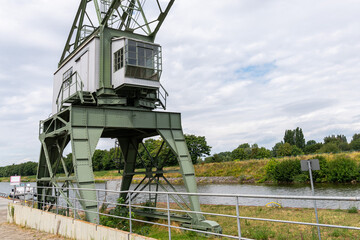 Part of a port crane with a cabin and a view of the river canal. A row of trees under a cloudy sky in the background.