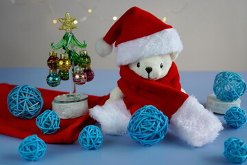 a white teddy bear in a red hat and Santa Claus scarf sits near a glass elegant Christmas tree with decorations in the form of colorful balls on a table on a gray background with garlands of bokeh