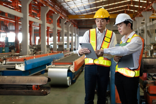 Industrial engineer and colleague wear safety helmet examining production in heavy steel engineering factory. Professional inspector and foreman worker inspecting material in manufacturing facility.