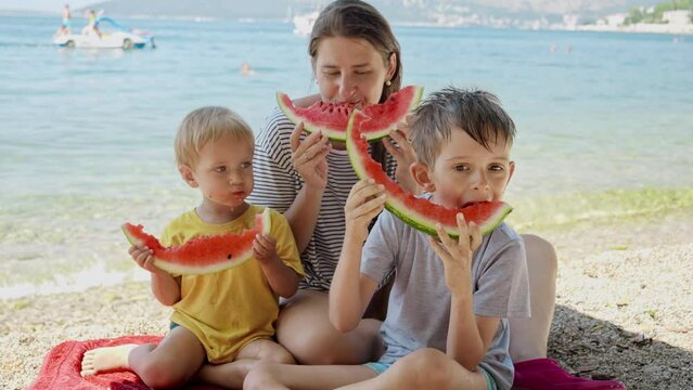 Mother and her two boys, captured in slow motion as they enjoy sweet watermelon on a beachside picnic. Family bonding during a holiday, summertime merriment, and the simple pleasures of a vacation.