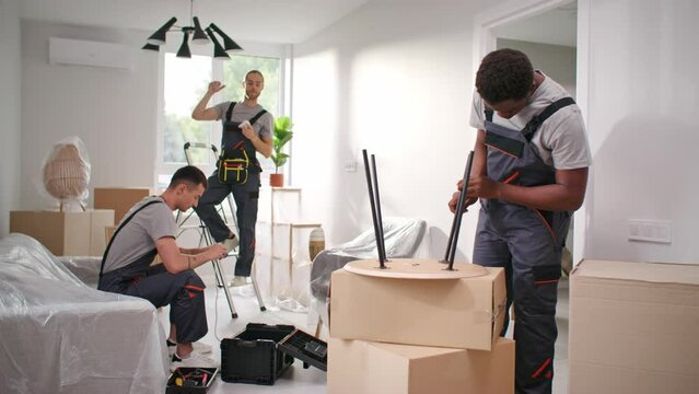 Three multiethnic men conducting repair work at apartment. Multicultural contractors making flat renovation. African American man screwing table legs. Electrician screwing light bulbs into chandelier.