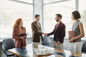 young architect and businessman shaking hands near colleagues and building model, closing deal