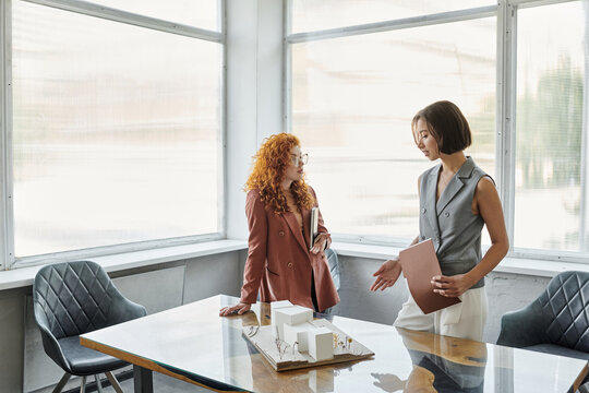 young businesswoman talking to redhead designer near architectural model in modern office