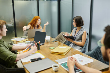 redhead businesswoman pointing with finger near colleagues, meeting room, laptops, documents, graphs