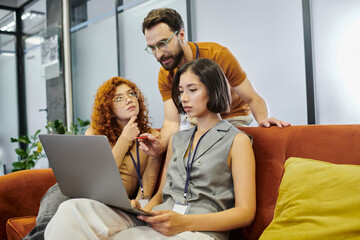 bearded team lead pointing at laptop near young women planning startup project in coworking office