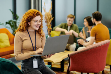 joyful redhead businesswoman with laptop near corporate team on blurred background in office