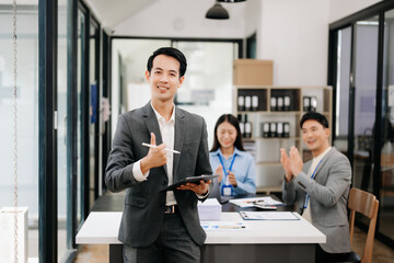 Young attractive Asian male office worker business suits smiling at camera with working notepad, tablet and laptop documents in office