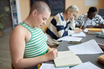 Side view of serious young woman sitting by desk and thinking of answer to one of test questions before writing it down in copybook