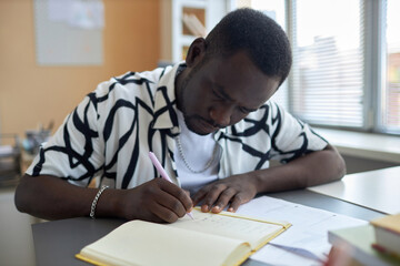Naklejka premium Serious African American male student bending over open copybook while sitting by desk at lesson and writing down answers to test questions