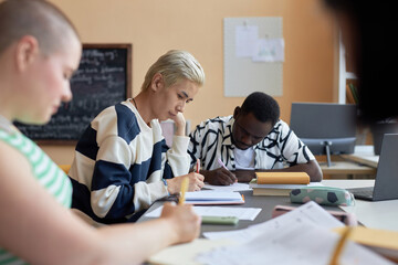 Fototapeta premium Focus on two young intercultural students carrying out examination test while sitting next to each other and writing down answers in copybooks