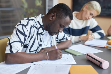 Naklejka premium Serious African American guy making notes or putting ticks on paper while answering questions of examination test against Asian classmate