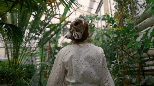 Back view of pretty young woman wearing white dress watching big leafs plants in tropical botanic garden