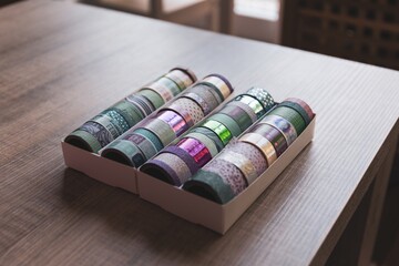 Naklejka premium A box full of colorful rolls of washi tape having different patterns, textures and colors on a wooden table. The tapes are ready to be used to decorate a post or gift card, an interior, a gift.