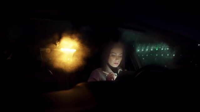 Young business woman sitting in the car during a rainy night browsing her electronic device