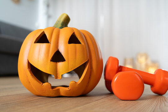 Halloween pumpkin ceramic decoration. Jack-o'-lantern spooky figurine and two dumbbells. Healthy fitness lifestyle, seasonal autumn or fall composition. Gym workout and sport training concept.