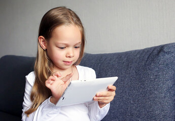 A little girl plays games on a tablet, holds it in her hands and looks at it.
