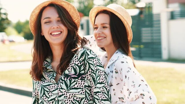 Two young beautiful smiling hipster women in trendy summer clothes. Carefree models posing on street background. Positive models having fun. Cheerful and happy, at sunny day in hats