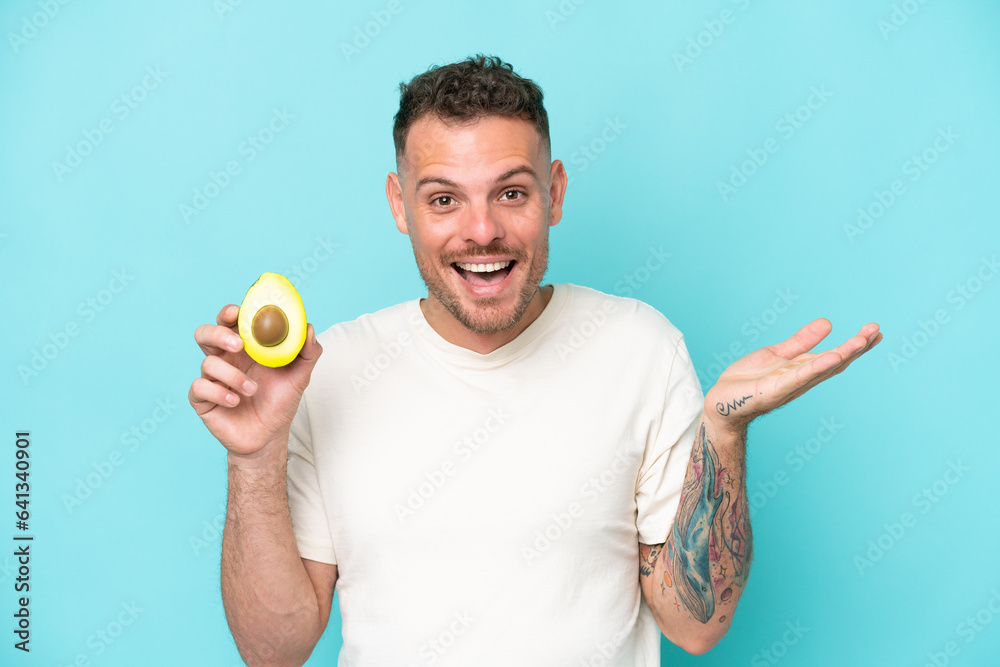 Wall mural young caucasian man holding an avocado isolated on blue background with shocked facial expression