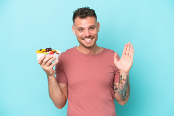 Young caucasian man holding a bowl of fruit isolated on blue background saluting with hand with happy expression