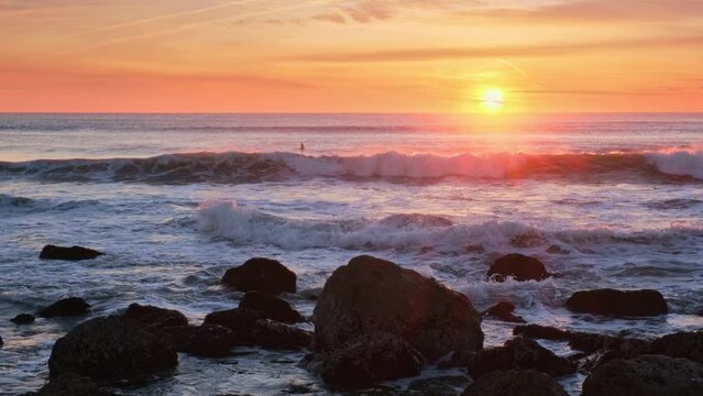 Atlantic ocean sunset with waves and rocks and surfers silhouettes in water at Costa da Caparica, Portugal