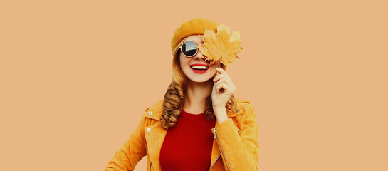 Autumn color style outfit, portrait of beautiful smiling young woman with yellow maple leaves wearing orange french beret hat, jacket on brown studio background - obrazy, fototapety, plakaty