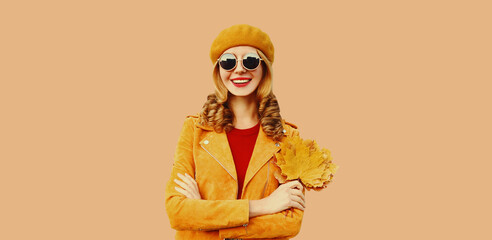 Autumn color style outfit, portrait of beautiful smiling young woman with yellow maple leaves wearing orange french beret hat, jacket on brown studio background