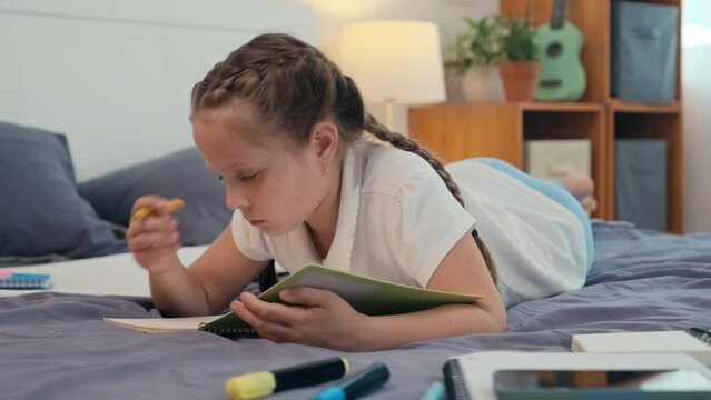 Pre-teen creative girl lying on bed while writing story in copybook, resting after schoolPre-teen creative girl lying on bed while writing story in copybook, resting after schoolPre-teen creative girl