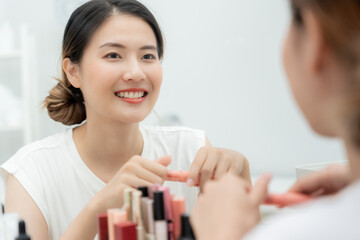 Beautiful Asian woman sit in front of a mirror and smile on makeup. face of a healthy woman applying makeup. Advertisement, lifestyle , cosmetics, makeup accessories, beauty activity, beautician