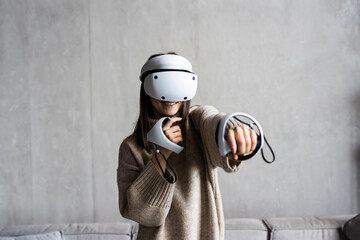 Young woman in vr goggles and with remote controllers playing video game while sitting on sofa in...