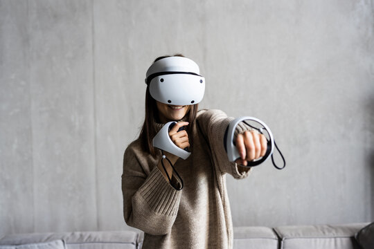Young woman in vr goggles and with remote controllers playing video game while sitting on sofa in living room