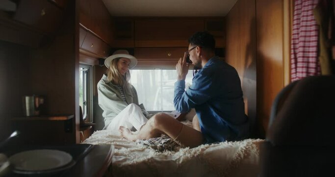 A man takes a picture of a woman in a hat sitting on a bed in a van. The woman smiles and poses