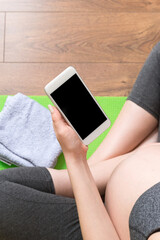 Pregnant female holding smartphone while sitting on exercise yoga mat at home at coronavirus time