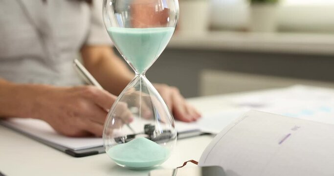 Hourglass measuring time on office desk business woman working over background. Efficiency deadlines and time management concept of busy employee