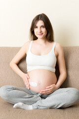 a pregnant girl sits at home on the bed and smears an anti-stretch mark cream on her stomach. Pregnancy, motherhood, preparation and expectation concept