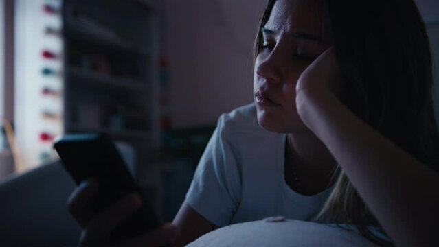 Depressed teenage girl lying on bed at home looking at bullying messages on mobile phone - shot in slow motion