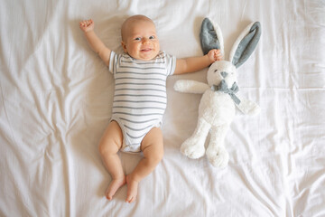 Adorable happy baby boy in bodysuit hugging toy and lying on back on bed at home room. Top view. 3...