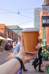 Fototapeta na wymiar White hand holding coffee paper cup on outside street market outside of St Lawrence Market in Toronto Canada