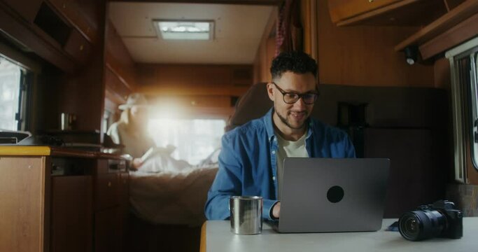 A young man uses a laptop and drinks a hot drink from a metal mug while sitting at a table in a van. A woman reads a book in the background