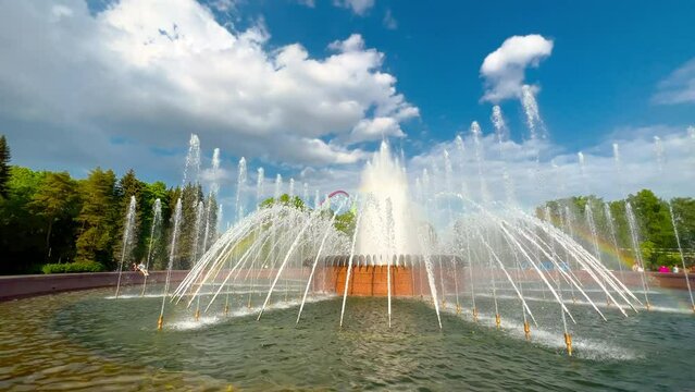 the huge fountain in city park, sunshine of splashes, sun reflections of water, rainbow, clear sunny weather, blue sky, green trees on background, no people