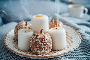 Fototapeta na wymiar Autumn fall cozy mood composition for hygge home decor. Orange pumpkins decorated with mandalas, white blown candles with smoke on wicker plate on the knitted gray plaid. Selective focus. Copy space